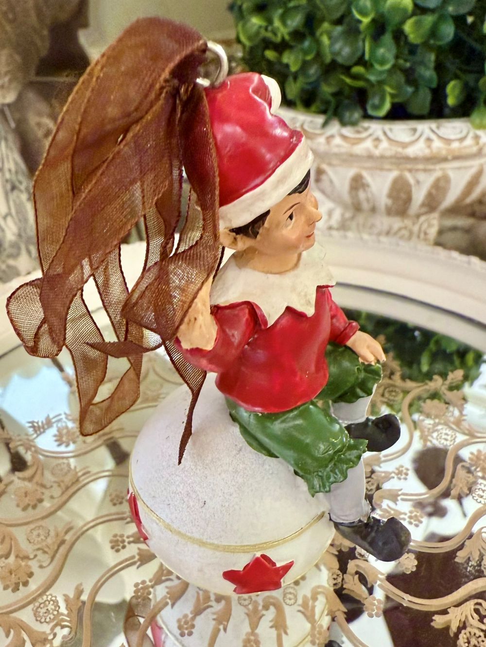 Suspension de Noël représentant un lutin rouge et vert assis sur une boule blanche décorée d’étoiles rouges, à suspendre dans le sapin, style féérique et romantique