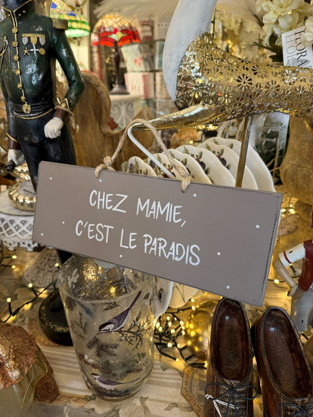 Plaque décorative en bois taupe avec inscription blanche « Chez Mamie c’est le paradis », style shabby chic, suspendue par une corde naturelle dans une ambiance romantique et chaleureuse
