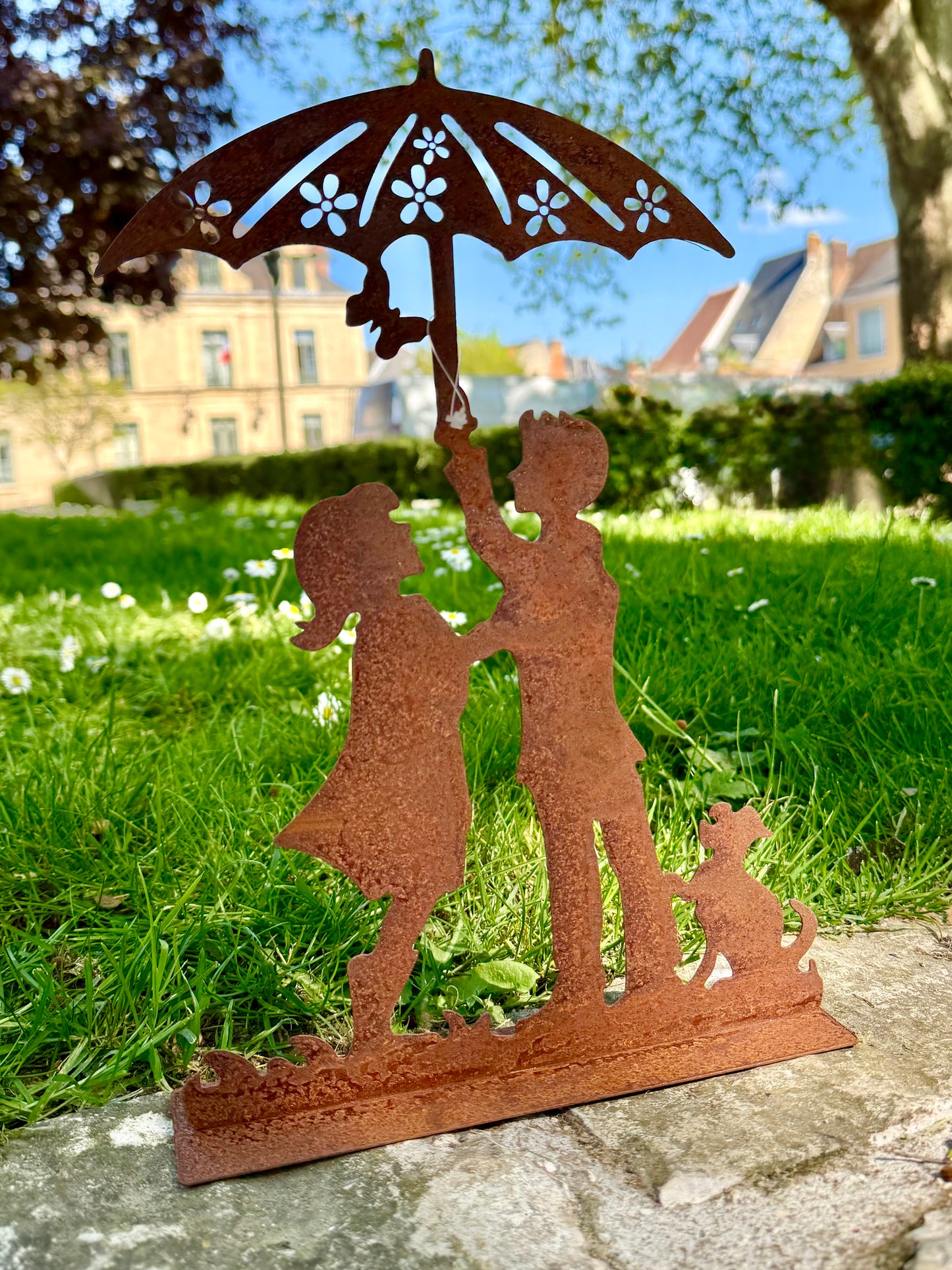 Silhouette décorative en fer forgé rouille représentant deux enfants sous une ombrelle avec un chien, posée sur une pelouse ensoleillée dans un jardin romantique.