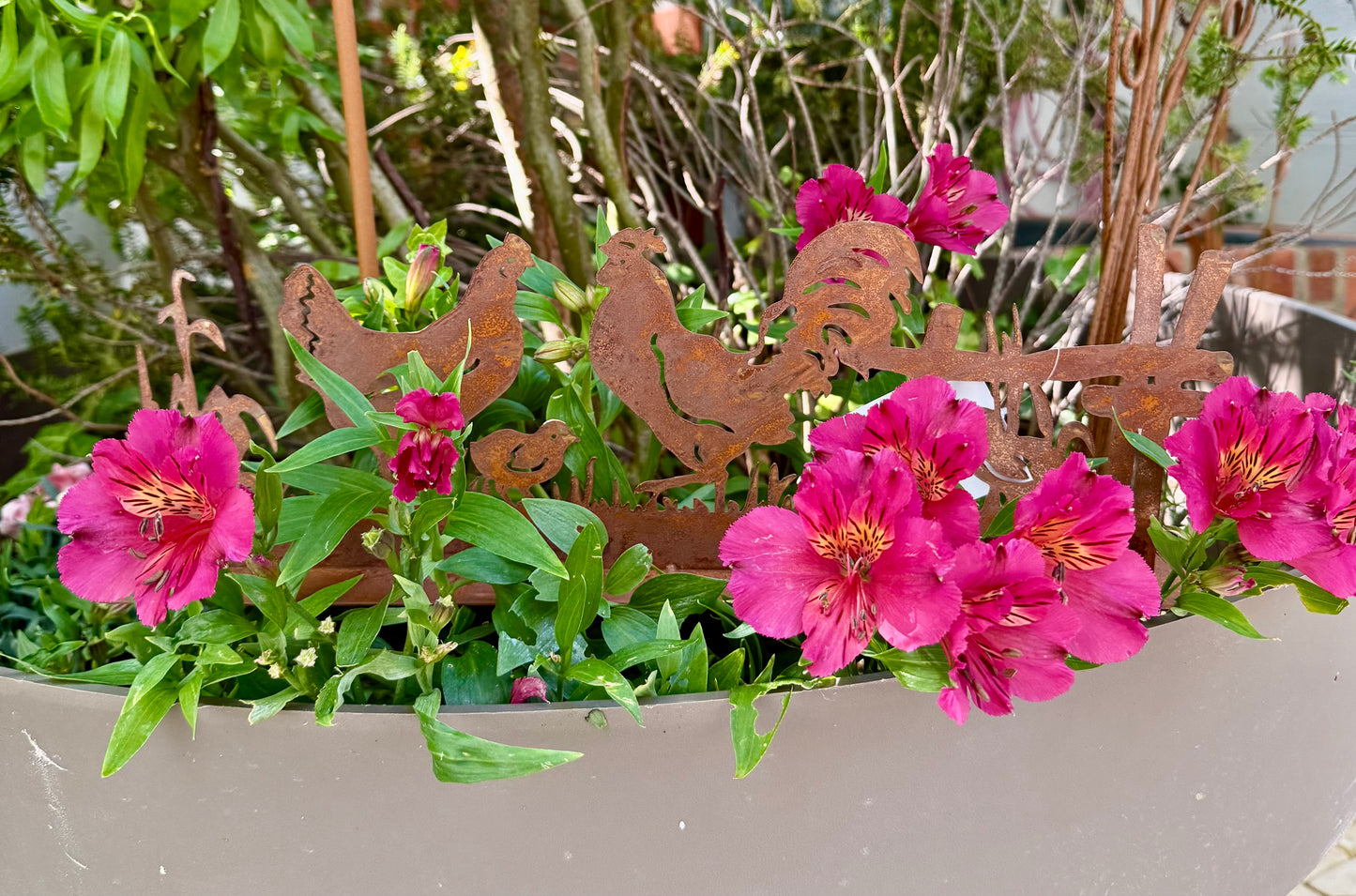 Silhouette décorative en fer forgé rouillé représentant une scène de basse-cour avec coq, poule et poussins, installée dans l’herbe d’un jardin champêtre