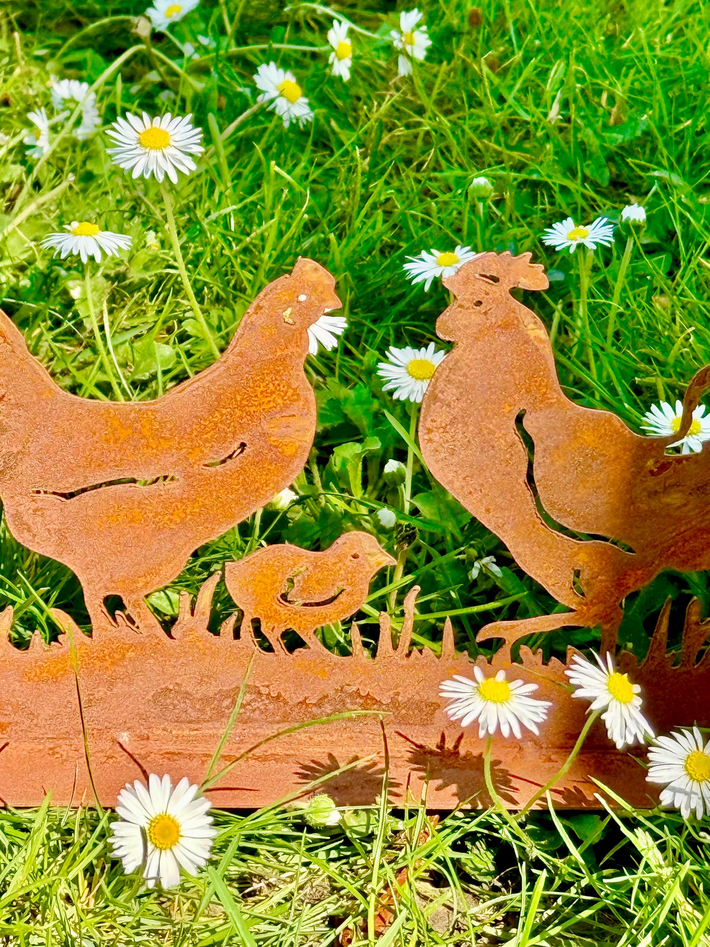 Silhouette décorative en fer forgé rouillé représentant une scène de basse-cour avec coq, poule et poussins, installée dans l’herbe d’un jardin champêtre