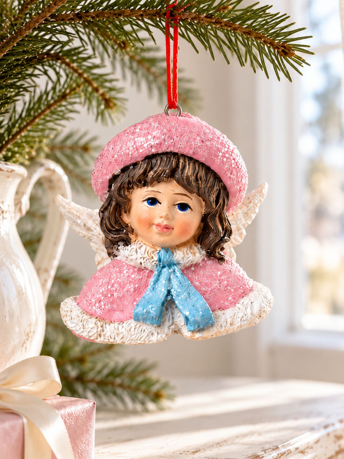 Ange Poupée Victorienne suspendu à un rameau de sapin dans un pichet blanc, près d’un cadeau rose.