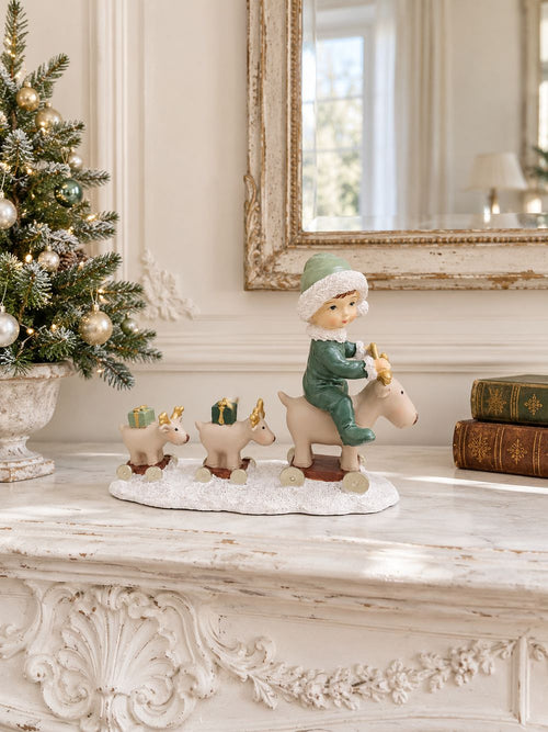 Statuette Balade des Petits Rennes sur un manteau de cheminée blanc patiné, entre un petit sapin décoré et des livres anciens.