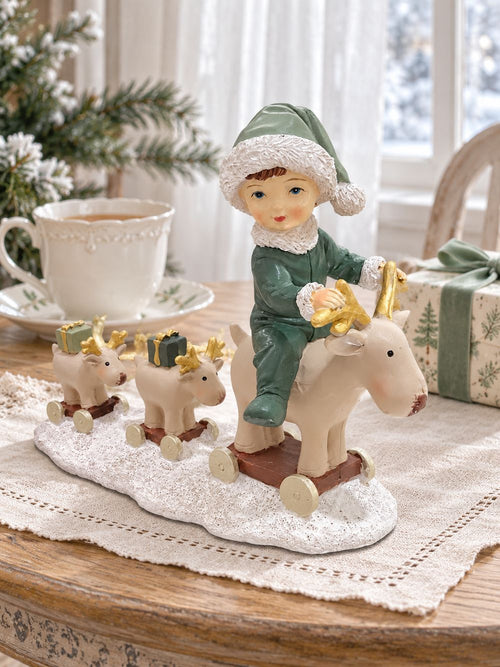 Statuette Balade des Petits Rennes sur une table près d’une fenêtre, accompagnée d’une tasse et d’un cadeau au ruban vert.