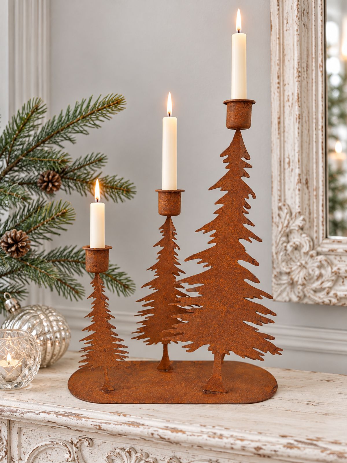Bougeoir à trois sapins en métal rouille avec bougies ivoire allumées, sur une console patinée blanche.