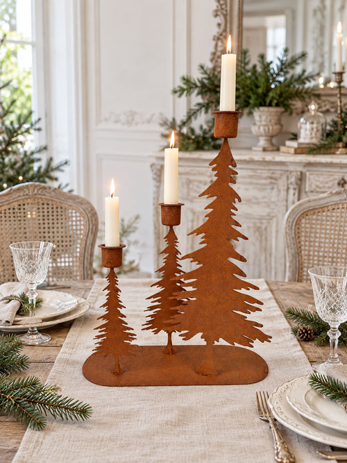 Bougeoir à trois sapins avec bougies allumées au centre d’une table de Noël, entre vaisselle ancienne et branches de sapin.