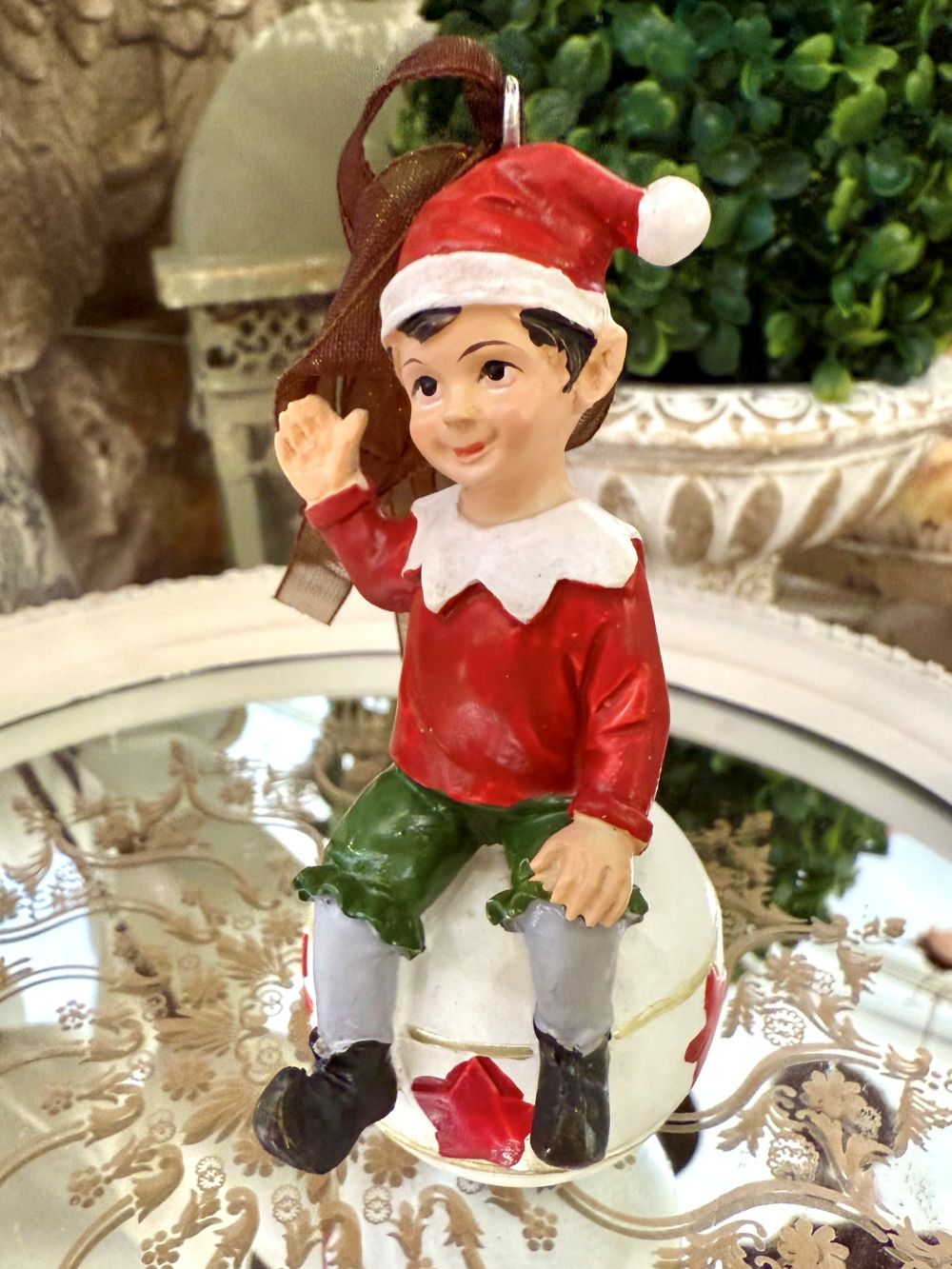 Suspension de Noël représentant un lutin rouge et vert assis sur une boule blanche décorée d’étoiles rouges, à suspendre dans le sapin, style féérique et romantique