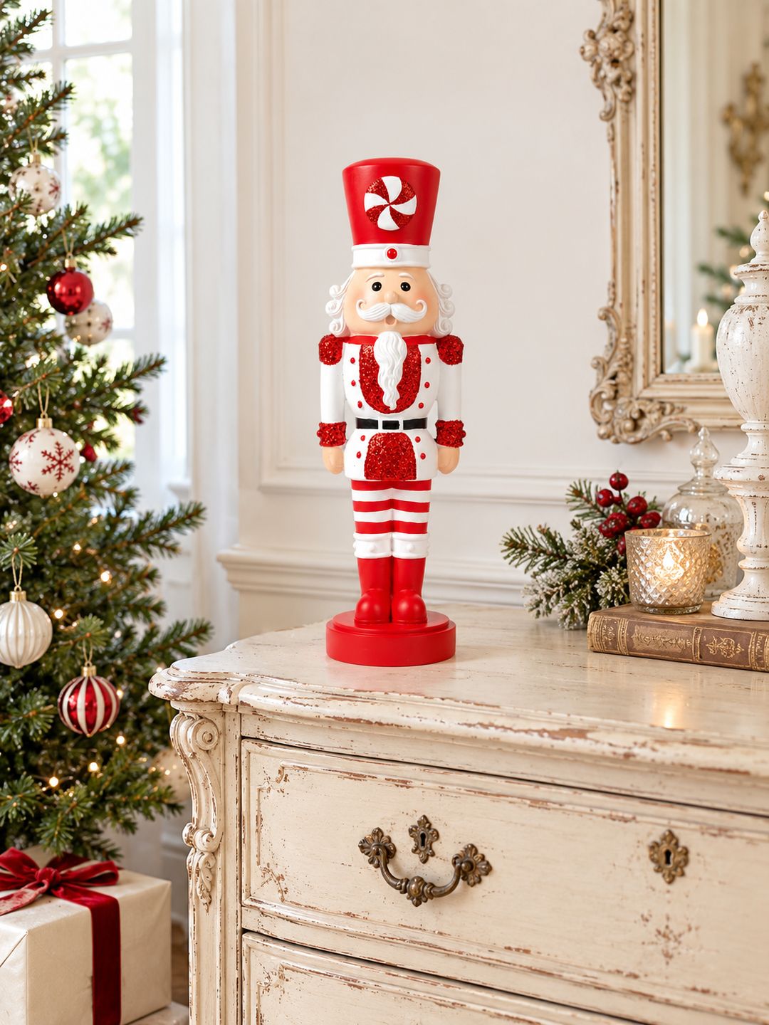 Casse-Noisette Candy Pailleté sur une commode ancienne crème, à côté d’un sapin décoré de boules rouges et blanches.