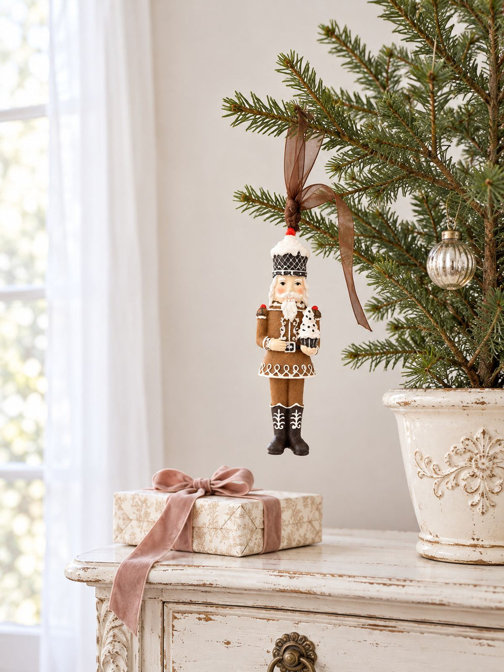 Casse-noisette Cupcake suspendu à un petit sapin sur une console patinée, près d’un cadeau au ruban rose.