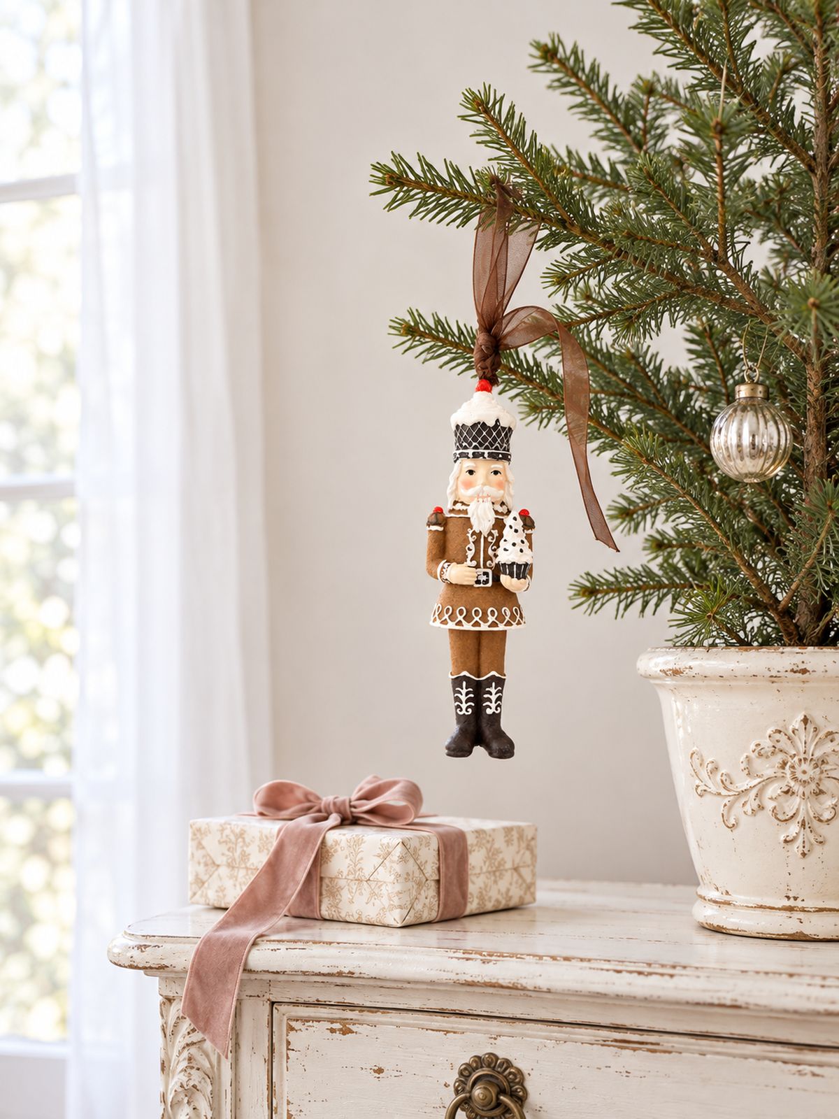 Casse-noisette Cupcake suspendu à un petit sapin sur une console patinée, près d’un cadeau au ruban rose.