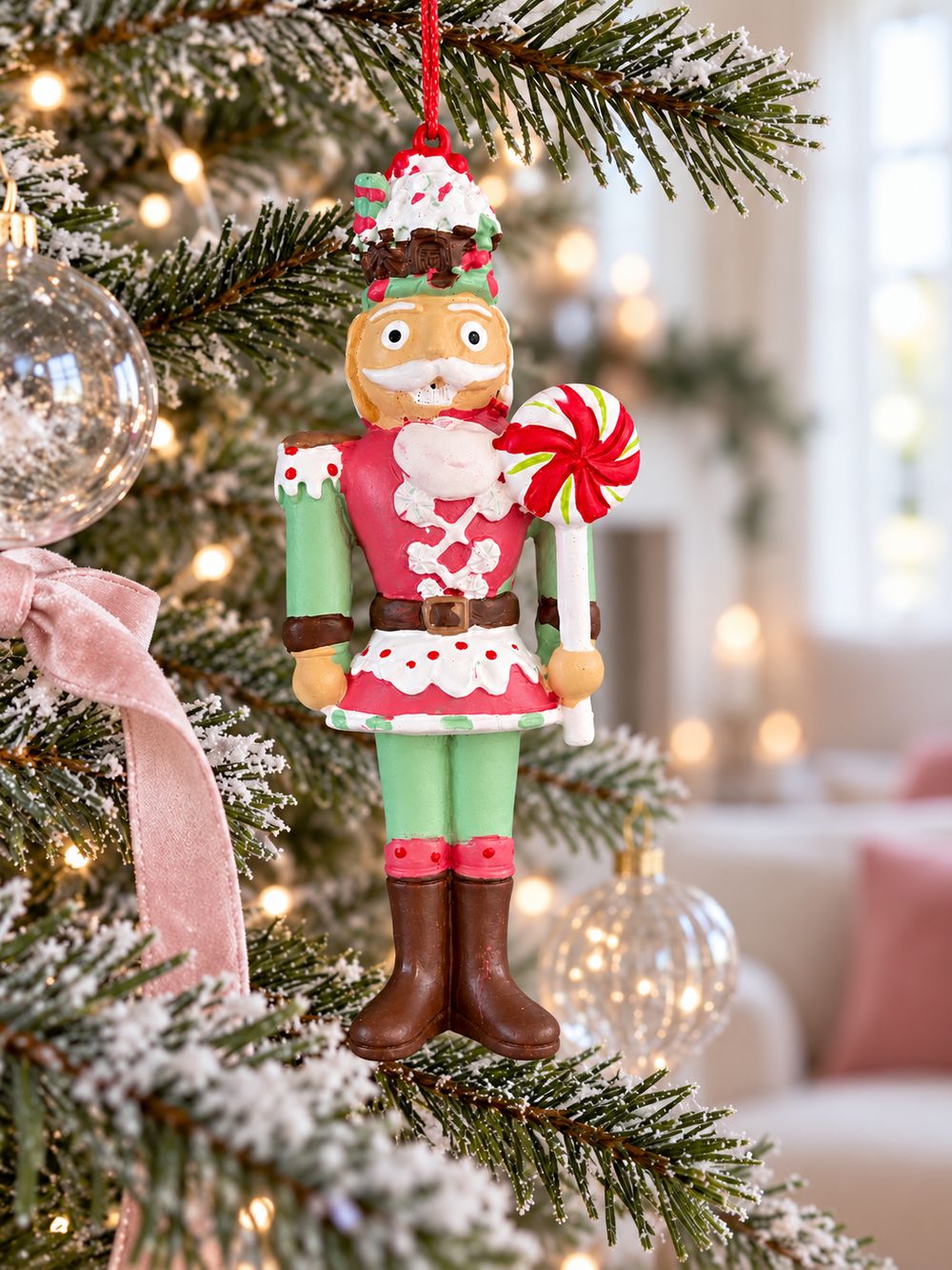 Casse-noisette suspendu verticalement dans un sapin de Noël, entouré de boules transparentes et d’un ruban rose poudré.