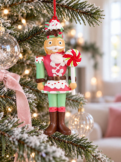 Casse-noisette suspendu verticalement dans un sapin de Noël, entouré de boules transparentes et d’un ruban rose poudré.