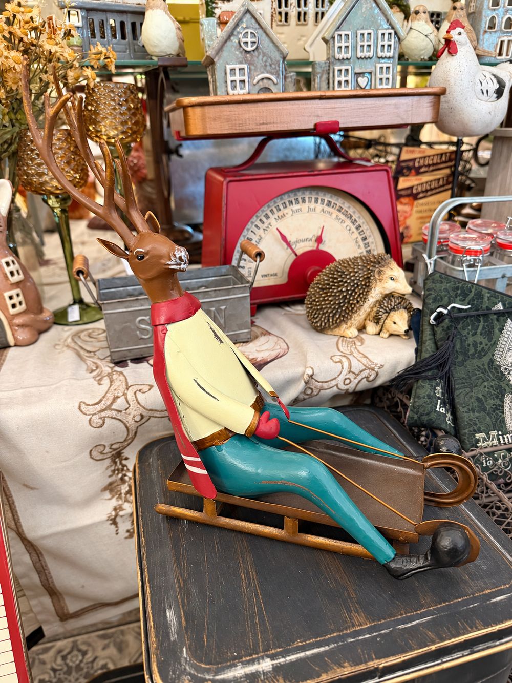 Cerf décoratif assis sur une luge, aux tons marron, rouge, jaune et bleu turquoise, style vintage et fantaisiste pour une décoration de Noël.