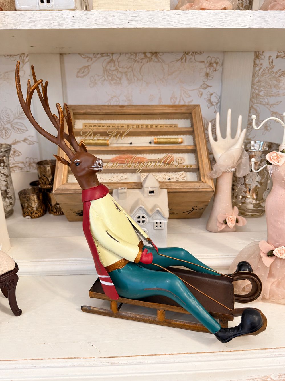 Cerf décoratif assis sur une luge, aux tons marron, rouge, jaune et bleu turquoise, style vintage et fantaisiste pour une décoration de Noël.