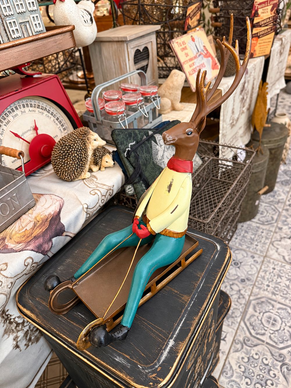 Cerf décoratif assis sur une luge, aux tons marron, rouge, jaune et bleu turquoise, style vintage et fantaisiste pour une décoration de Noël.