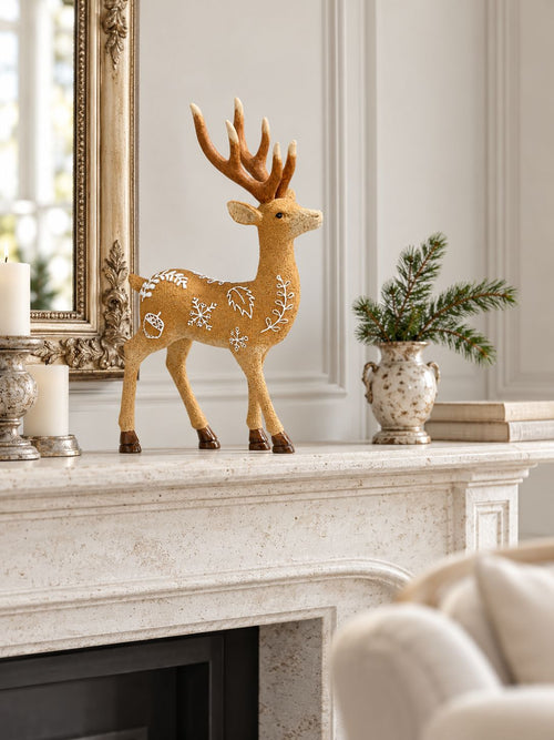Cerf décoratif Le Prince des Forêts sur une cheminée en pierre claire dans un salon lumineux, près d’un vase de branches de sapin.
