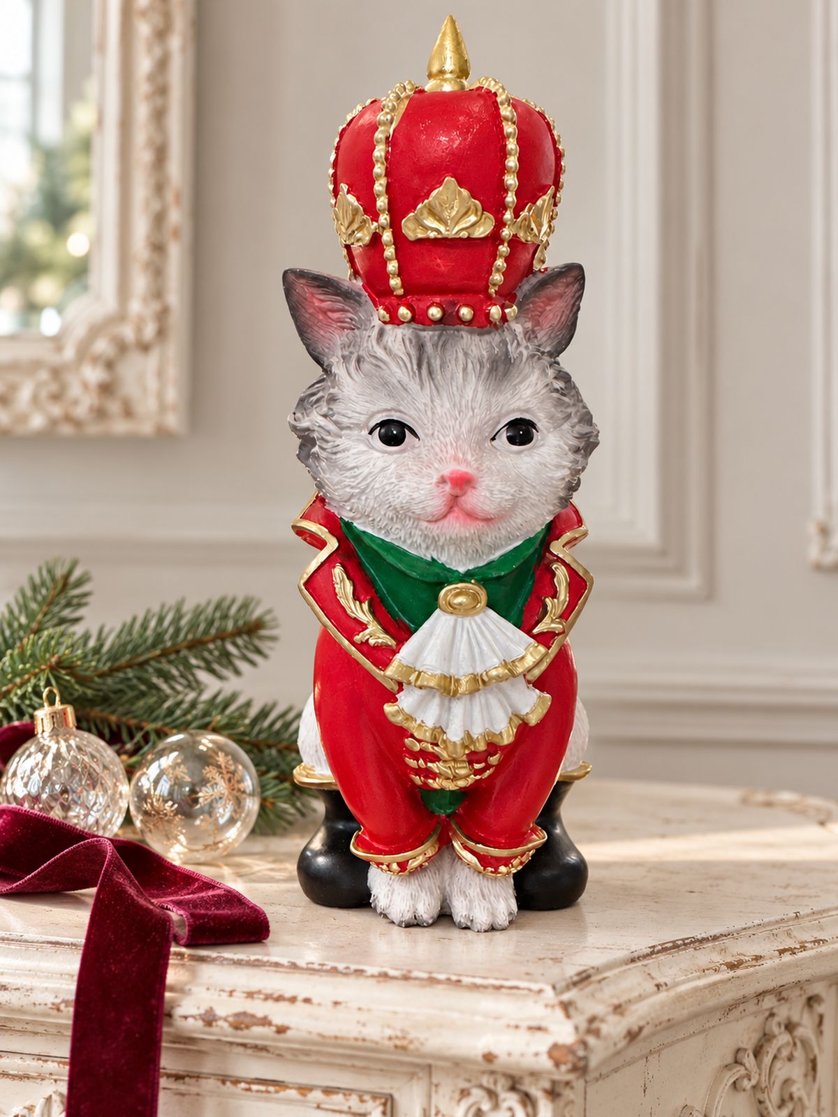 Figurine de chat couronné en costume rouge et or sur une console patinée, avec des boules de Noël et une branche de sapin.