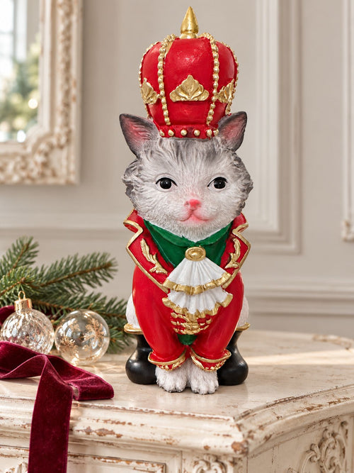 Figurine de chat couronné en costume rouge et or sur une console patinée, avec des boules de Noël et une branche de sapin.