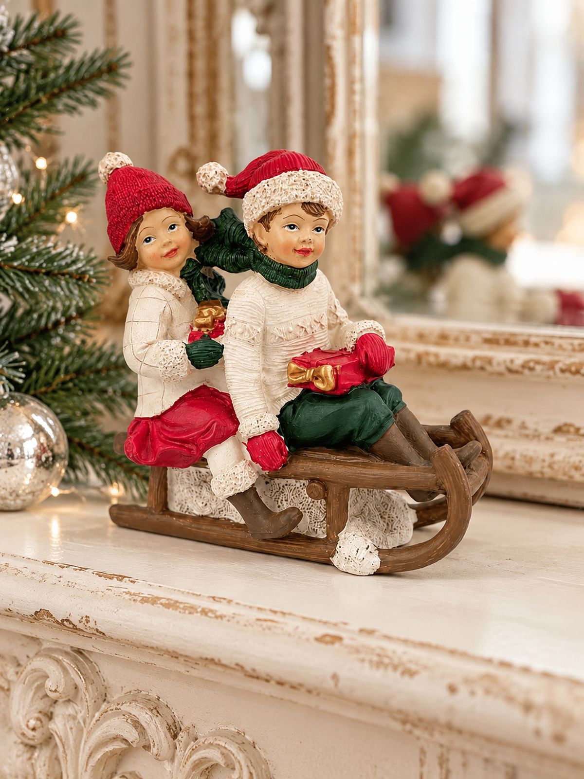 Figurine Chérubins sur une luge posée sur une cheminée patinée, devant un miroir et des branches de sapin.