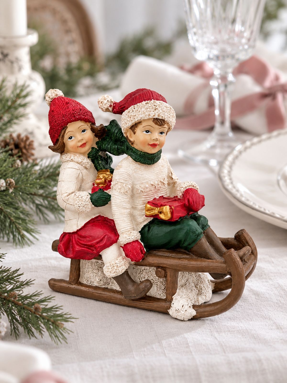 Figurine Chérubins sur une luge sur une table de Noël en lin blanc, près d’une assiette et d’un verre à pied.