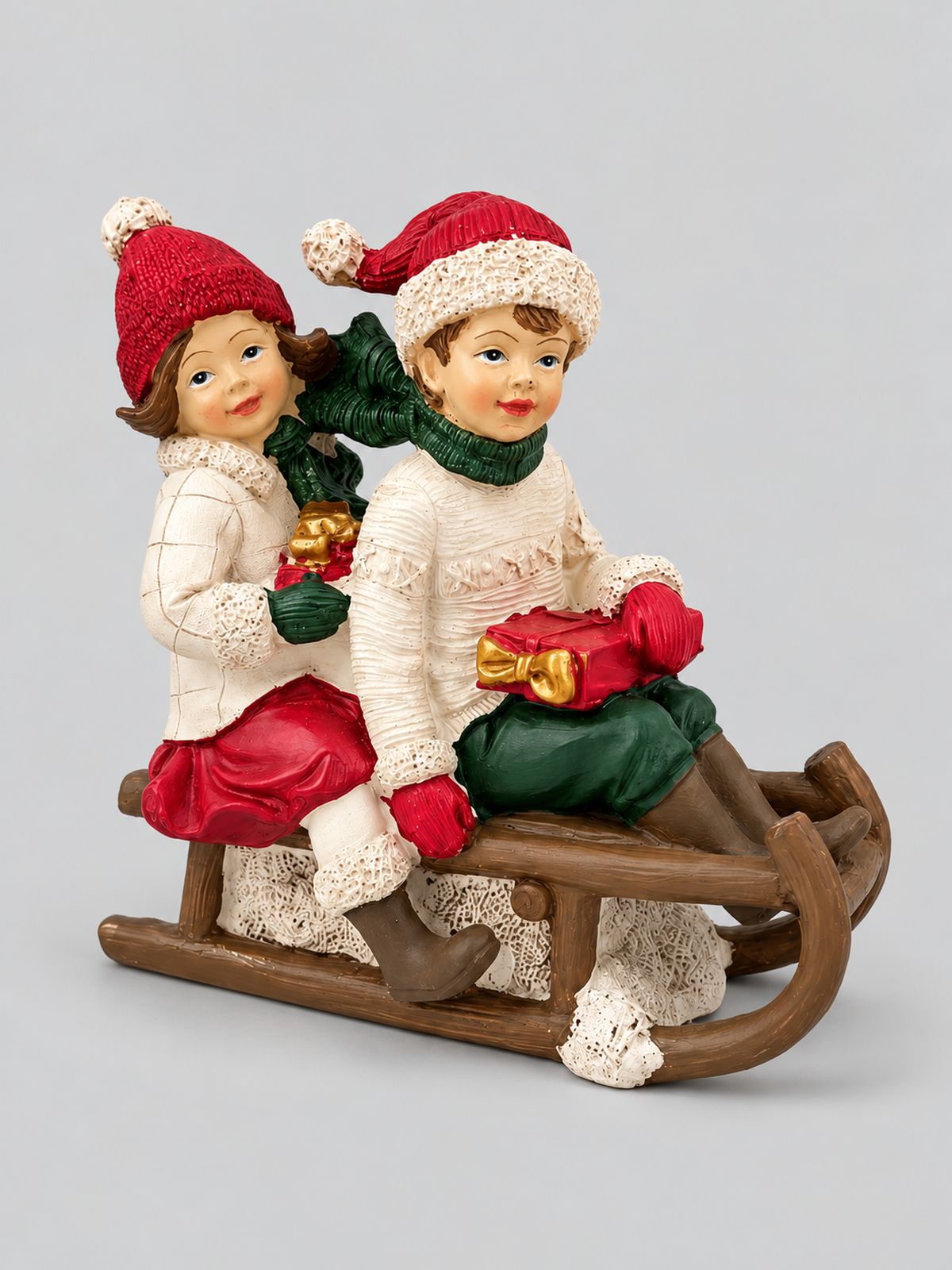 Figurine Chérubins sur une luge, avec bonnets rouges et cadeaux, présentée sur fond gris clair.