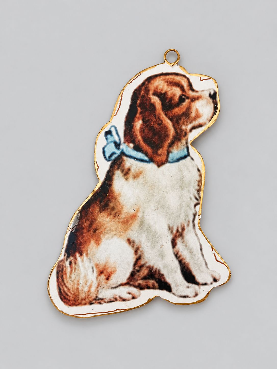 Suspension de Noël en métal représentant un chien victorien au nœud bleu, bordée de doré, sur fond gris clair.