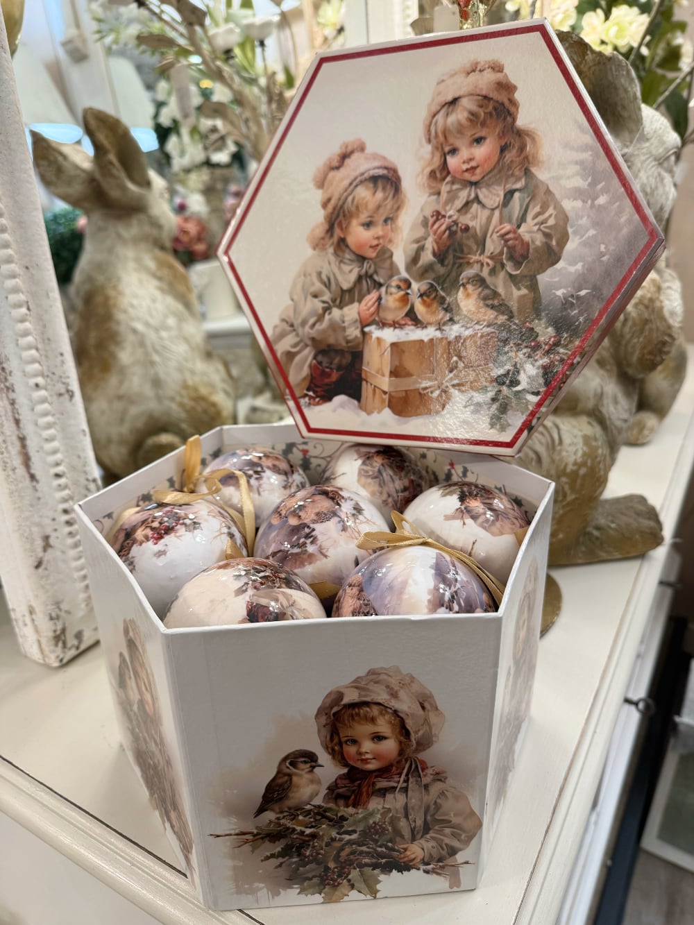 Coffret de boules de Noël décoratives illustrées de scènes d'enfance et de petits oiseaux, dans des tons crème et beige, suspendues sur un sapin enneigé au style romantique et vintage