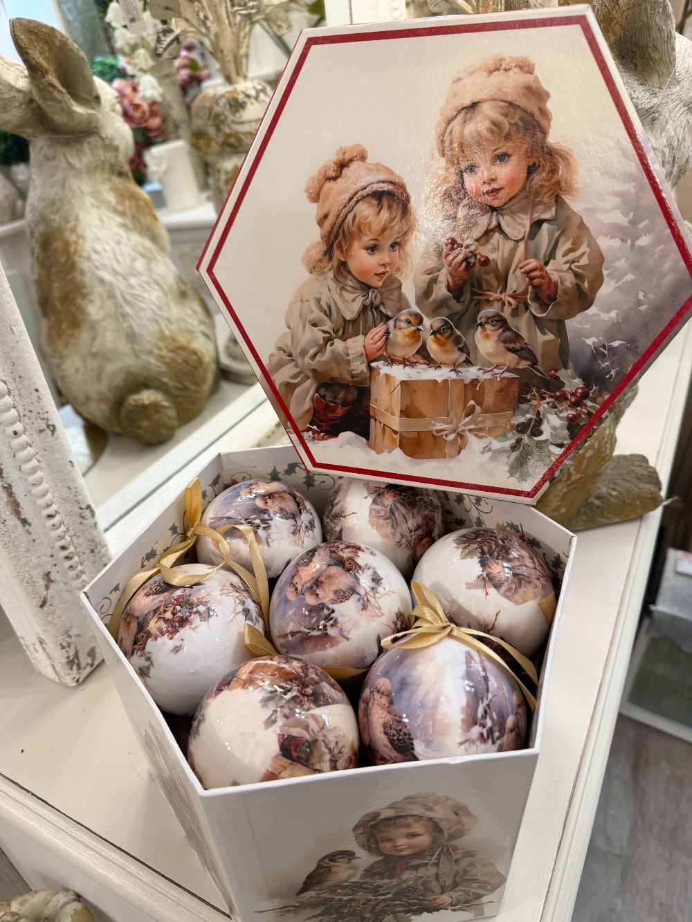 Coffret de boules de Noël décoratives illustrées de scènes d'enfance et de petits oiseaux, dans des tons crème et beige, suspendues sur un sapin enneigé au style romantique et vintage