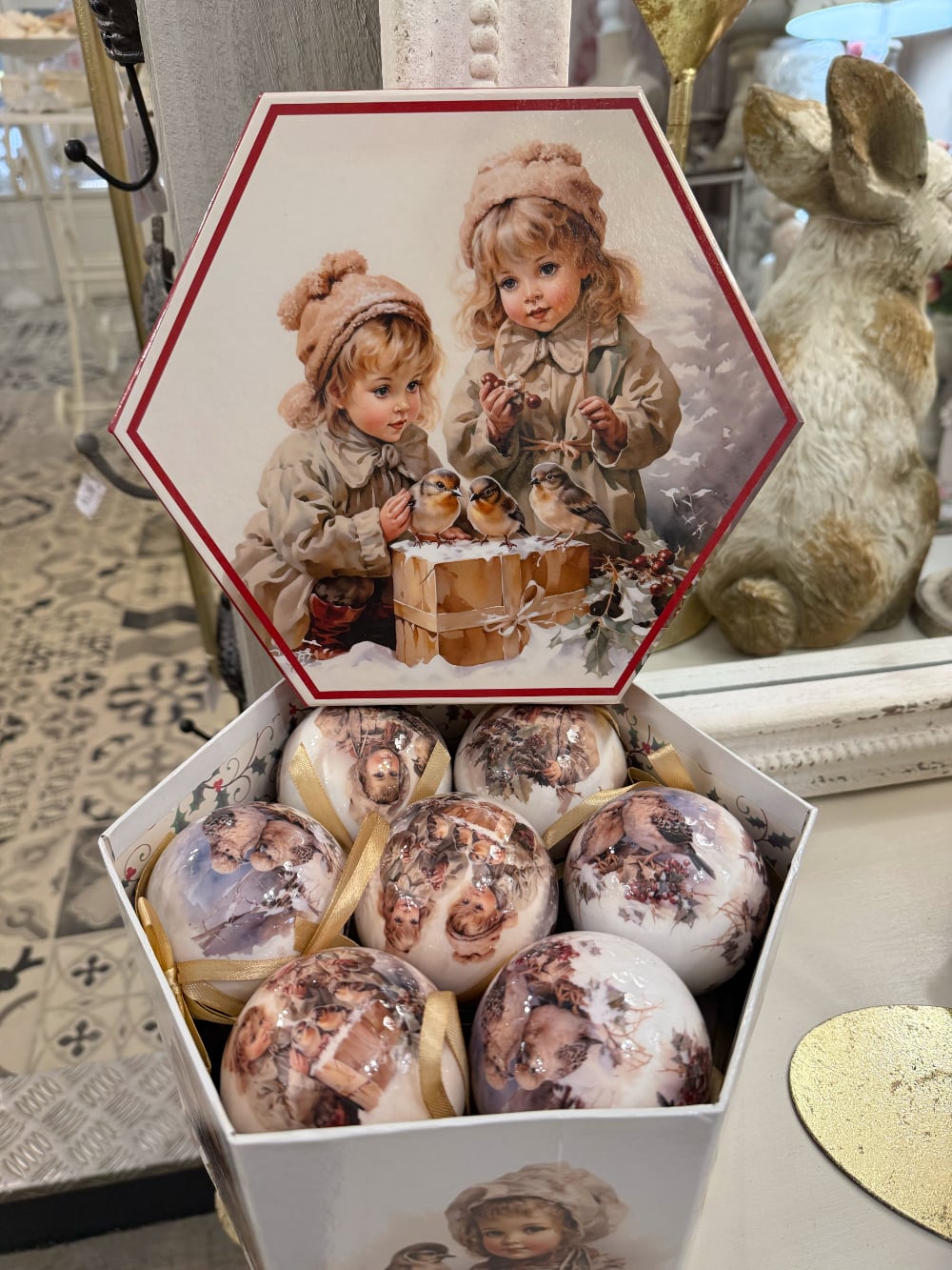 Coffret de boules de Noël décoratives illustrées de scènes d'enfance et de petits oiseaux, dans des tons crème et beige, suspendues sur un sapin enneigé au style romantique et vintage