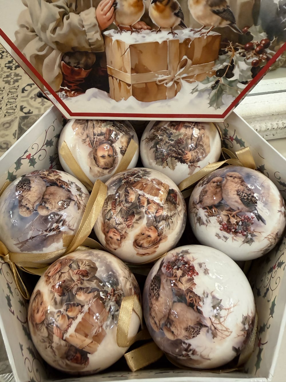 Coffret de boules de Noël décoratives illustrées de scènes d'enfance et de petits oiseaux, dans des tons crème et beige, suspendues sur un sapin enneigé au style romantique et vintage
