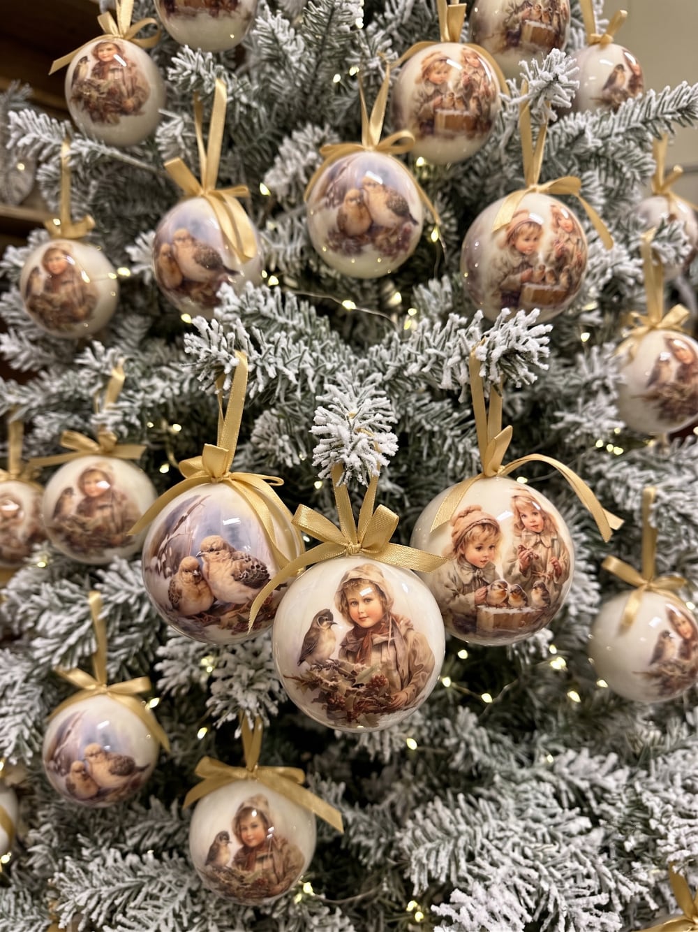 Coffret de boules de Noël décoratives illustrées de scènes d'enfance et de petits oiseaux, dans des tons crème et beige, suspendues sur un sapin enneigé au style romantique et vintage