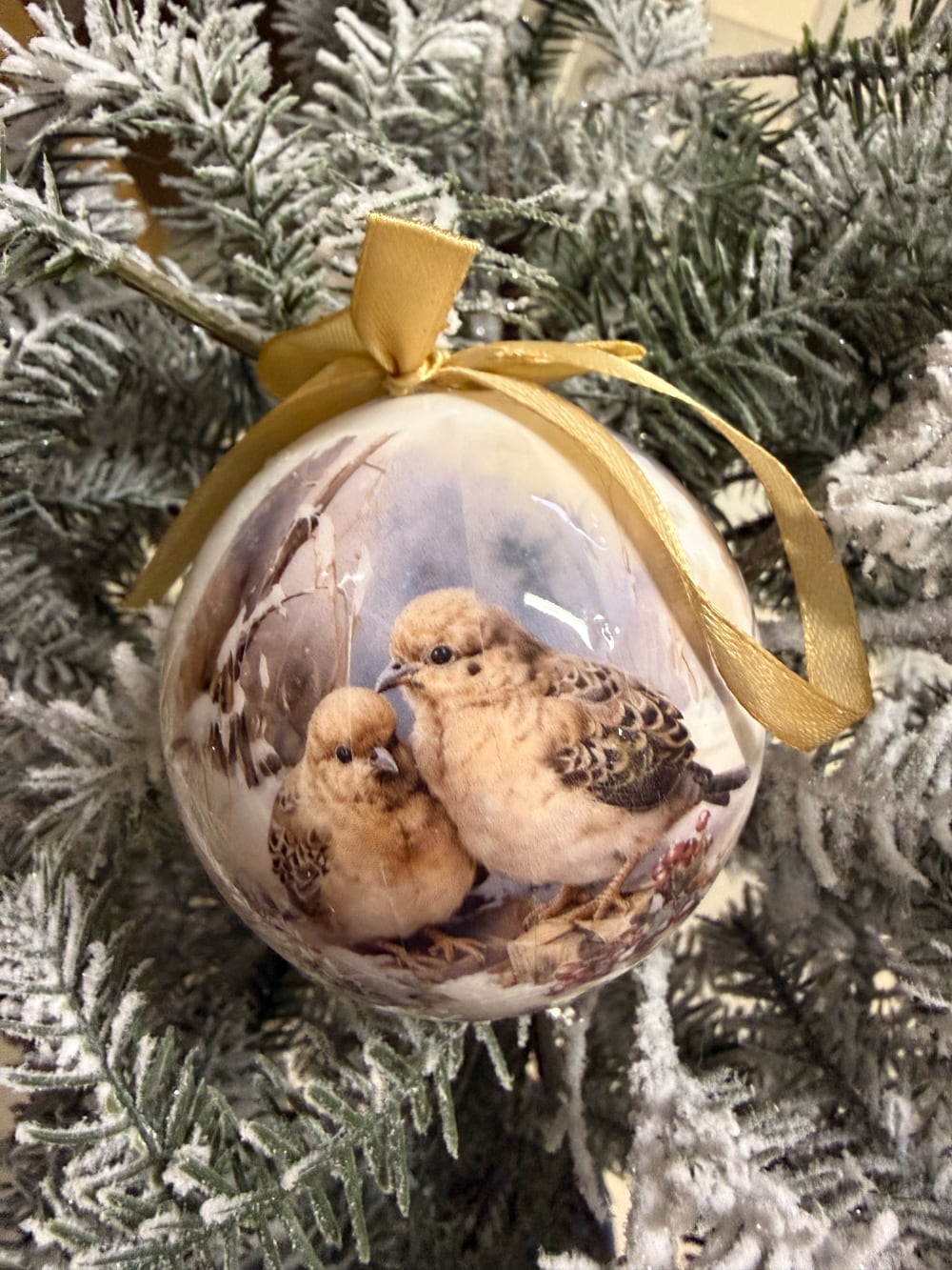 Coffret de boules de Noël décoratives illustrées de scènes d'enfance et de petits oiseaux, dans des tons crème et beige, suspendues sur un sapin enneigé au style romantique et vintage