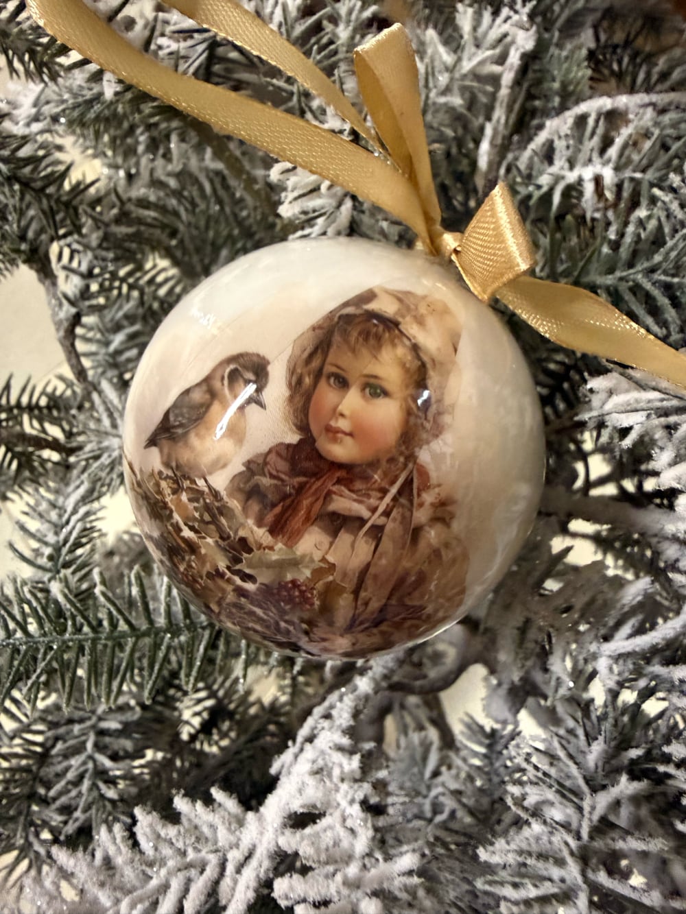 Coffret de boules de Noël décoratives illustrées de scènes d'enfance et de petits oiseaux, dans des tons crème et beige, suspendues sur un sapin enneigé au style romantique et vintage