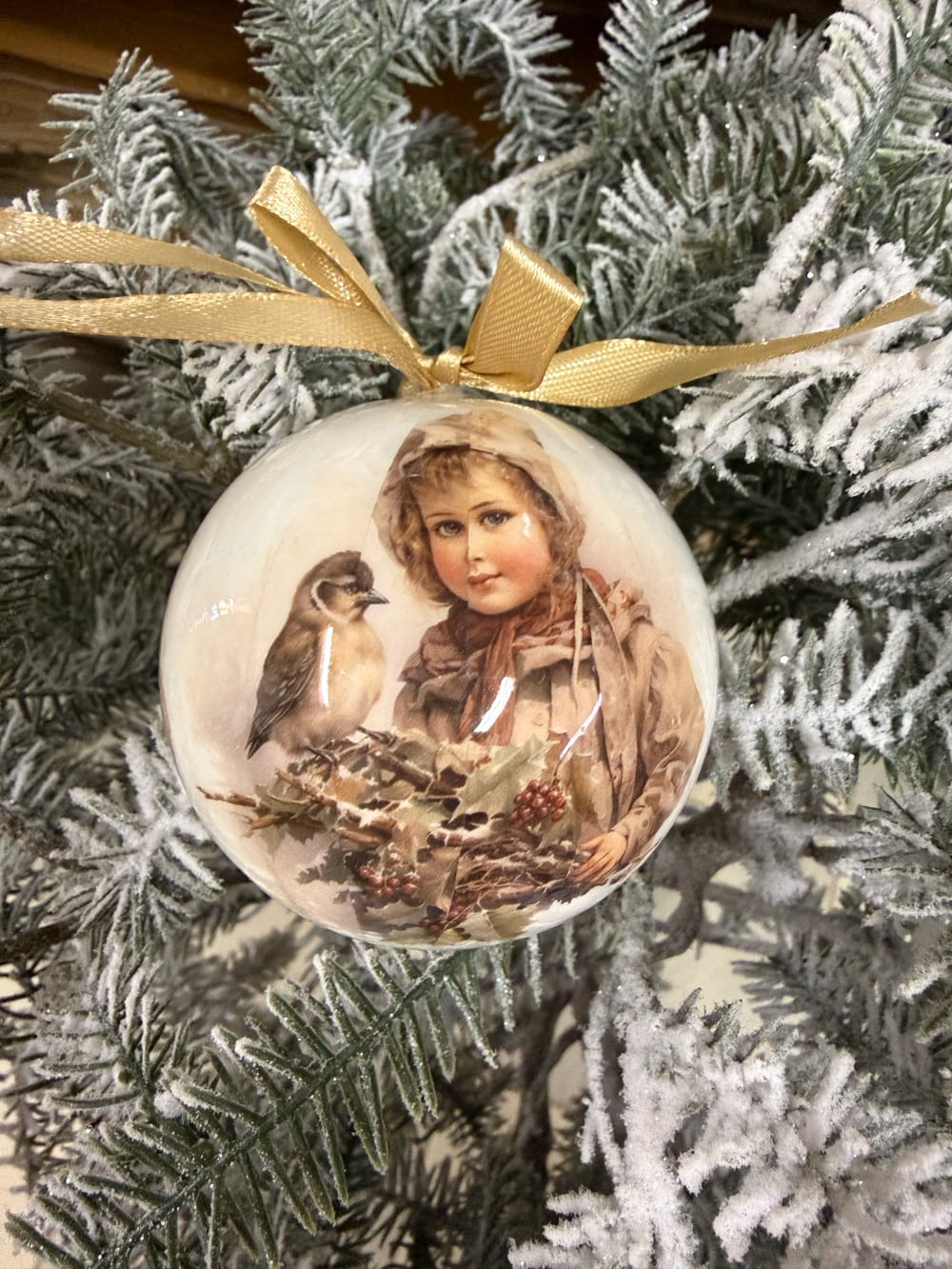 Coffret de boules de Noël décoratives illustrées de scènes d'enfance et de petits oiseaux, dans des tons crème et beige, suspendues sur un sapin enneigé au style romantique et vintage