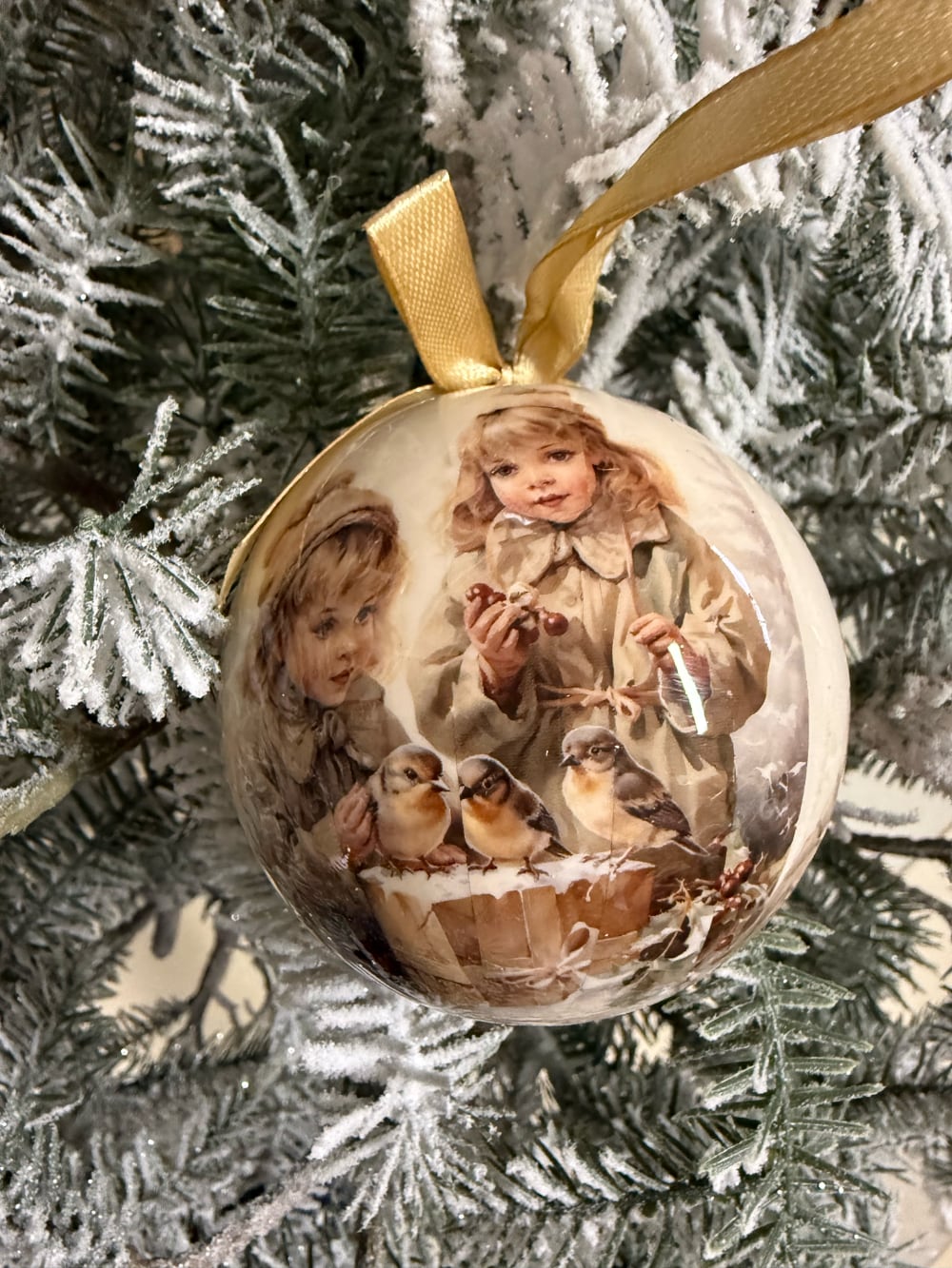Coffret de boules de Noël décoratives illustrées de scènes d'enfance et de petits oiseaux, dans des tons crème et beige, suspendues sur un sapin enneigé au style romantique et vintage
