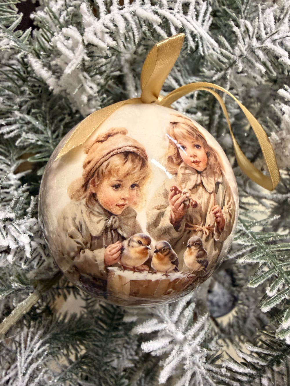 Coffret de boules de Noël décoratives illustrées de scènes d'enfance et de petits oiseaux, dans des tons crème et beige, suspendues sur un sapin enneigé au style romantique et vintage