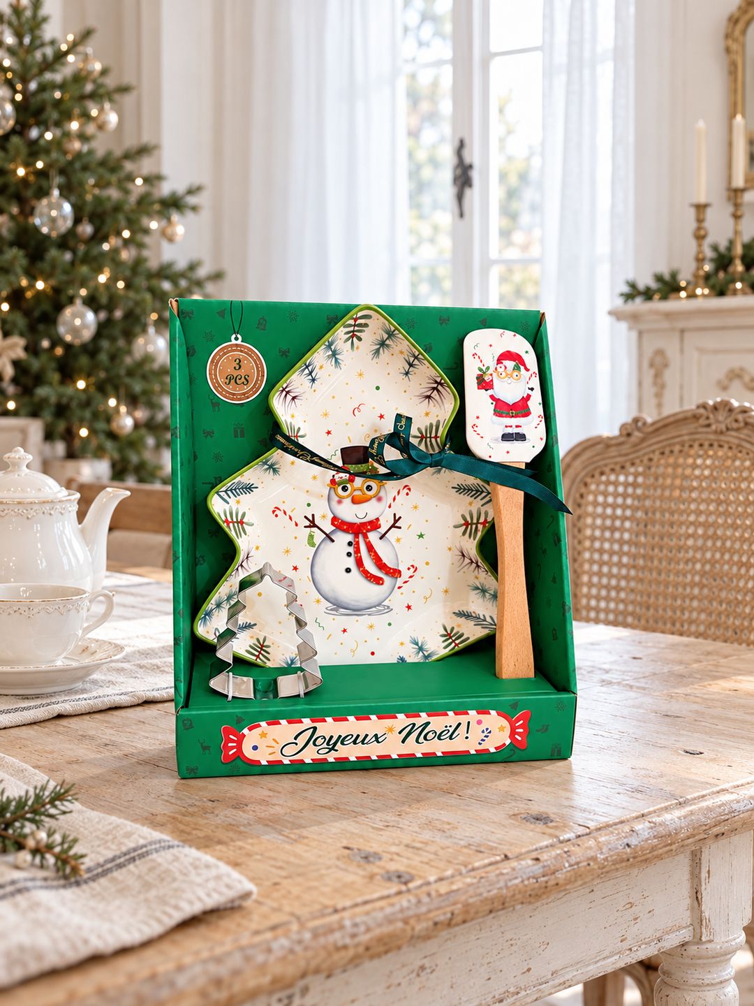 Coffret Snowman de Noël sur une table ancienne avec un service à thé, devant un sapin dans une salle à manger lumineuse.