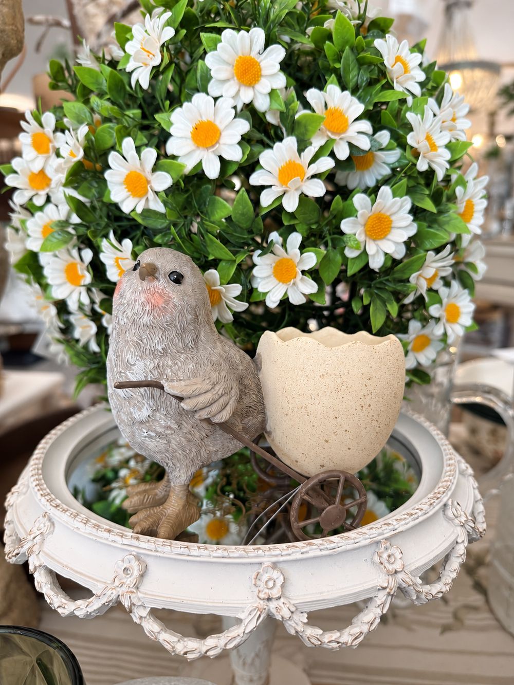 Coquetier poussin et sa coquille d’œuf sur roues, présentés sur un plateau miroir devant des marguerites.