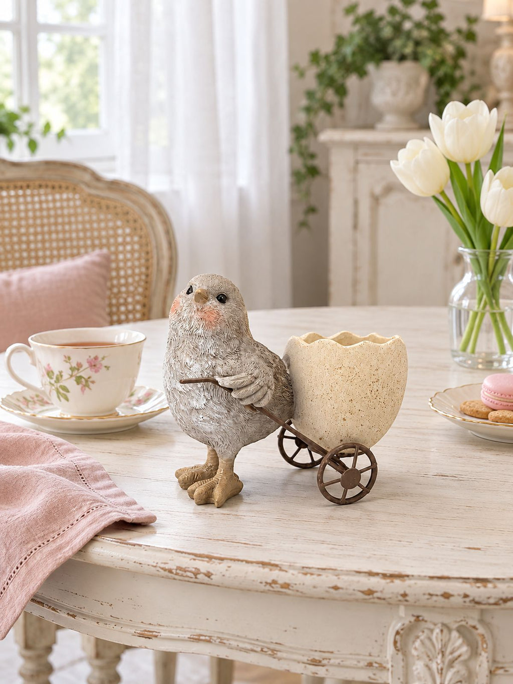 Coquetier poussin sur une table ronde patinée, avec tasse fleurie et tulipes blanches dans un intérieur lumineux.