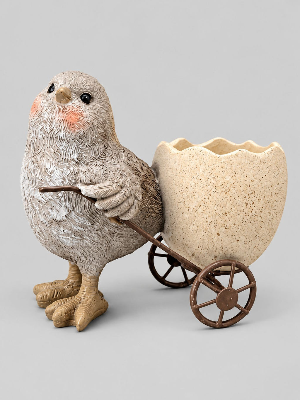 Coquetier poussin gris aux joues roses tirant une coquille d’œuf crème sur roues brunes, sur fond gris clair.