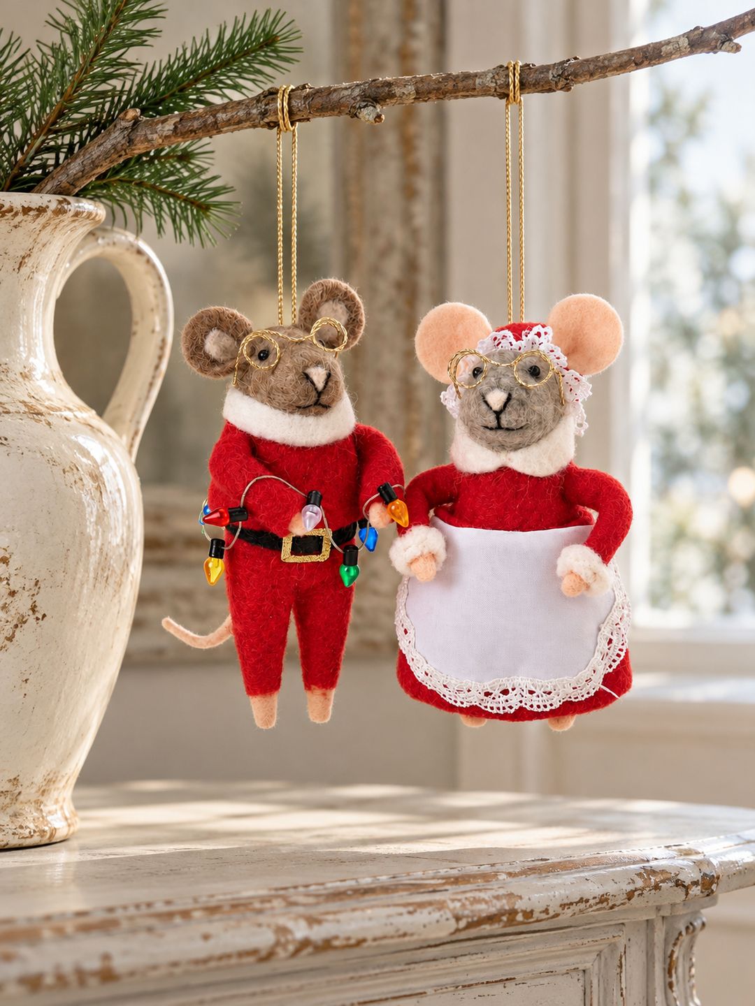 Deux souris de Noël en feutrine suspendues à une branche dans une cruche ancienne, au-dessus d’une console patinée.