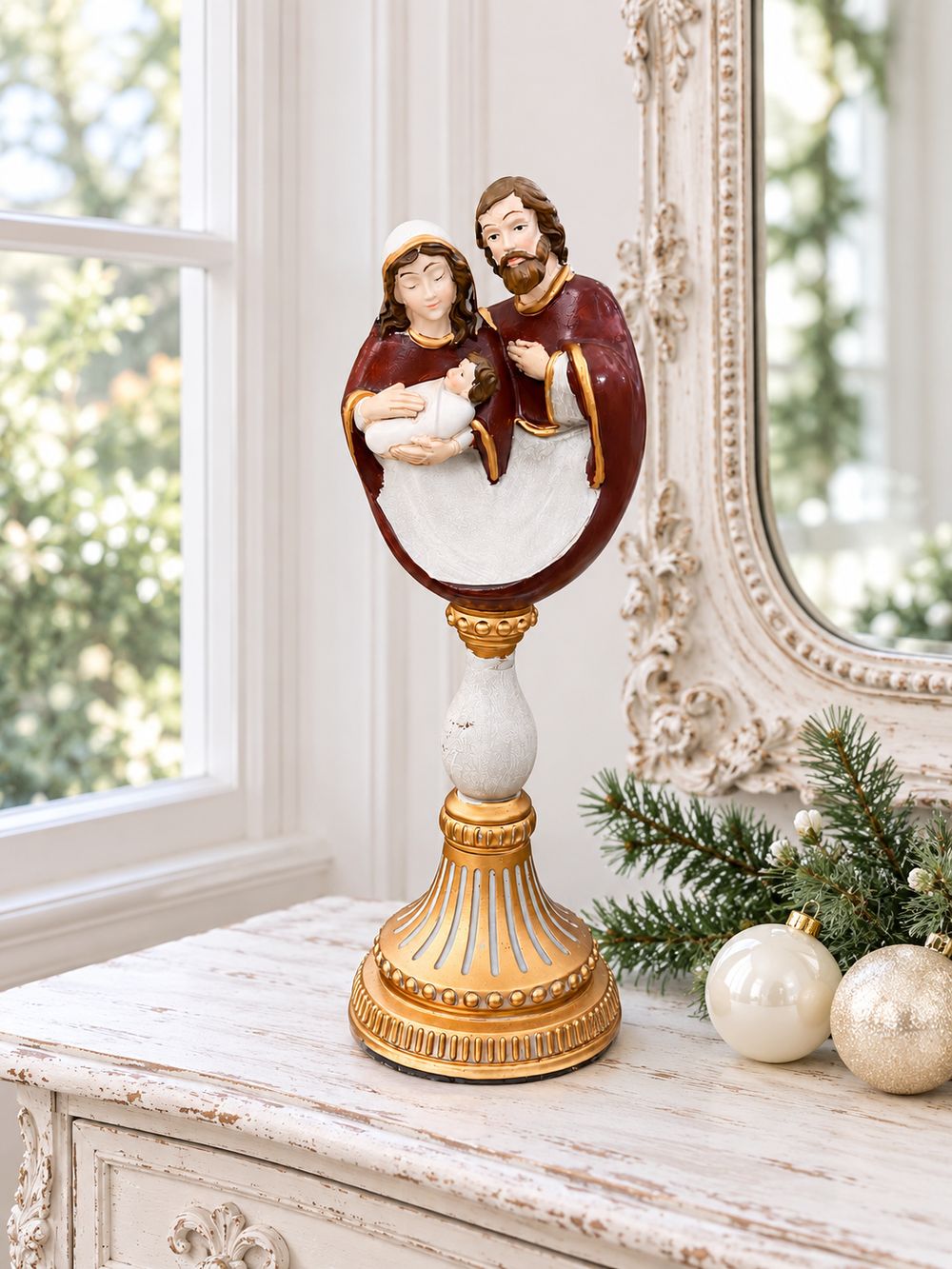 Crèche de la Sainte Famille sur socle doré, posée sur une console patinée près d’une fenêtre et de décorations de Noël nacrées.
