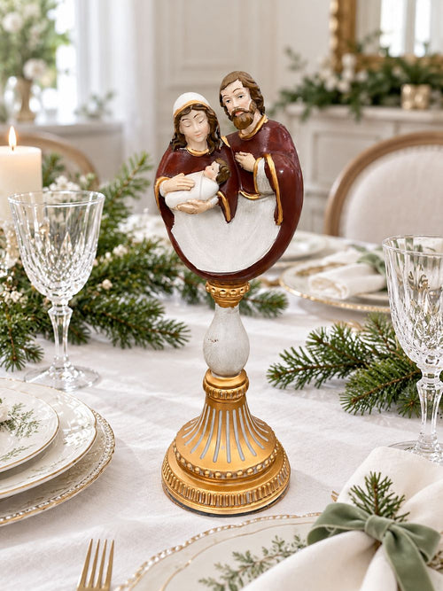 Crèche sur socle baroque doré au centre d’une table de Noël, entourée de porcelaine, de verres à pied et de branches de sapin.