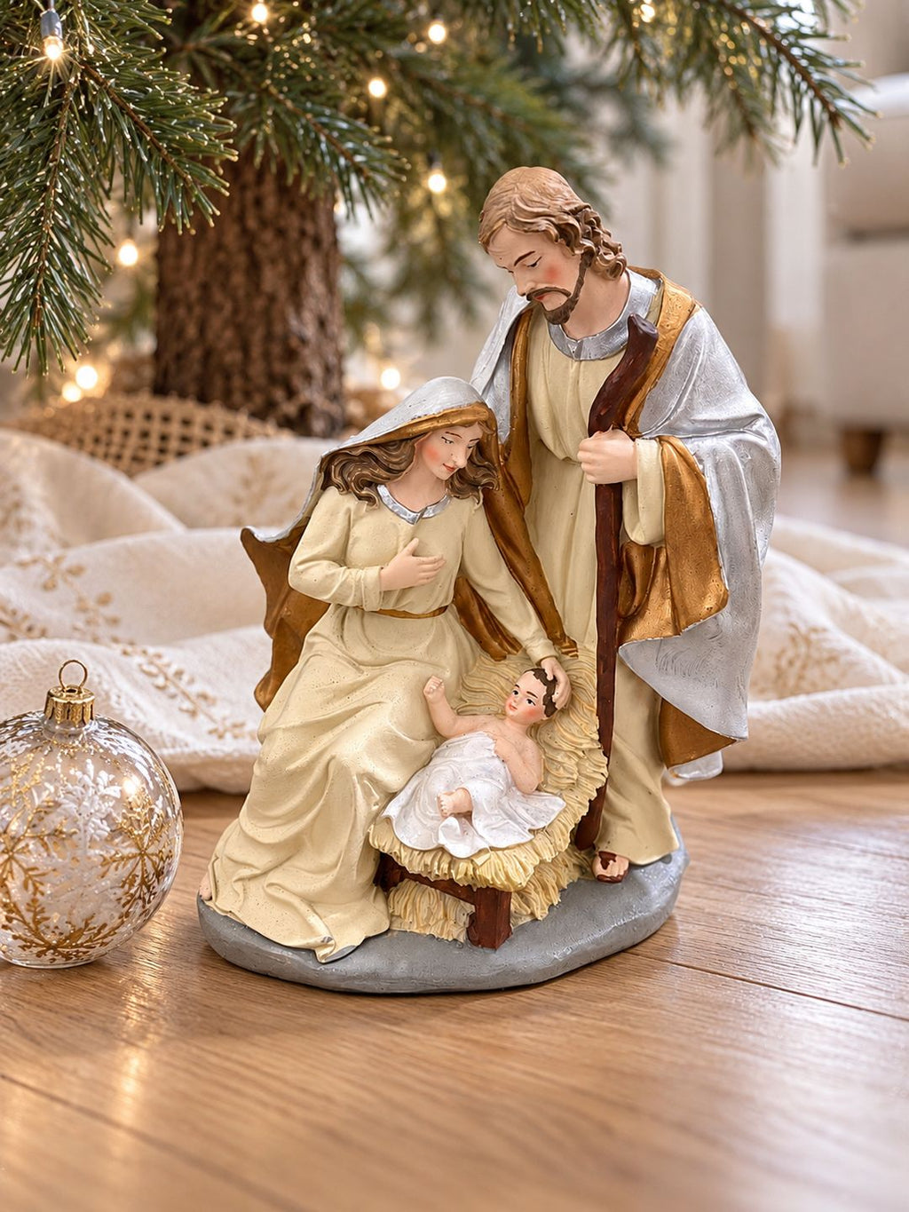 Crèche de Noël en résine crème, dorée et argentée, posée au pied d’un sapin sur un parquet clair, près d’une boule de Noël en verre.