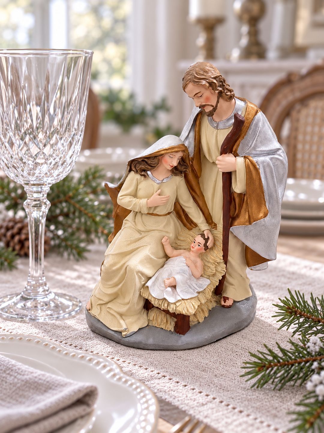 Crèche de Noël sur une table claire, près d’un verre à pied, de vaisselle blanche et de branches de sapin.