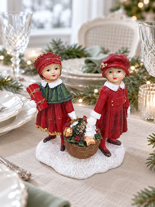 Figurine Enfants au Panier de Noël sur une table de fête en lin, entourée de vaisselle blanche et de verres.