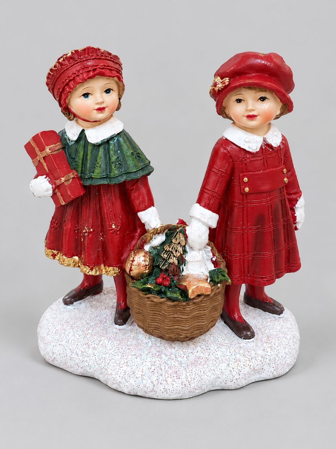 Figurine Enfants au Panier de Noël, deux enfants en tenues rouges et vertes sur un socle enneigé, sur fond gris clair.