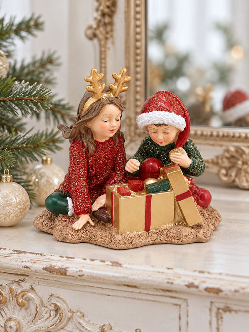 Figurine Les Enfants au Trésor de Noël sur une console blanche patinée, devant un miroir ancien, avec des branches de sapin et des boules ivoire.