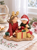 Figurine Les Enfants au Trésor de Noël sur une table au linge brodé, accompagnée d’une tasse fleurie et de sablés étoilés.