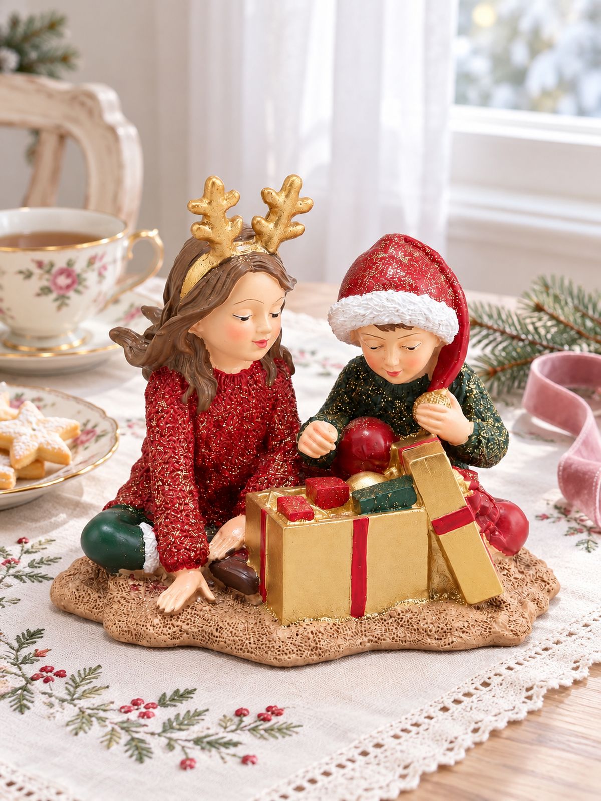 Figurine Les Enfants au Trésor de Noël sur une table au linge brodé, accompagnée d’une tasse fleurie et de sablés étoilés.